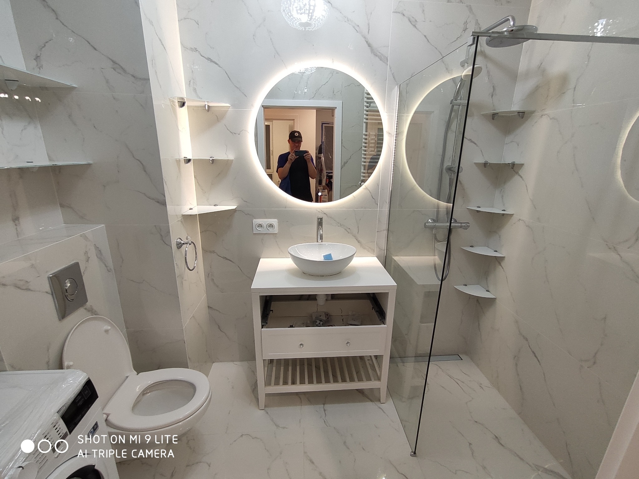 Nowoczesna łazienka z białymi marmurkowymi płytkami, okrągłym lustrem z podświetleniem LED, kabiną prysznicową typu walk-in i umywalką nablatową na białej szafce; widoczne półki szklane i toaleta.