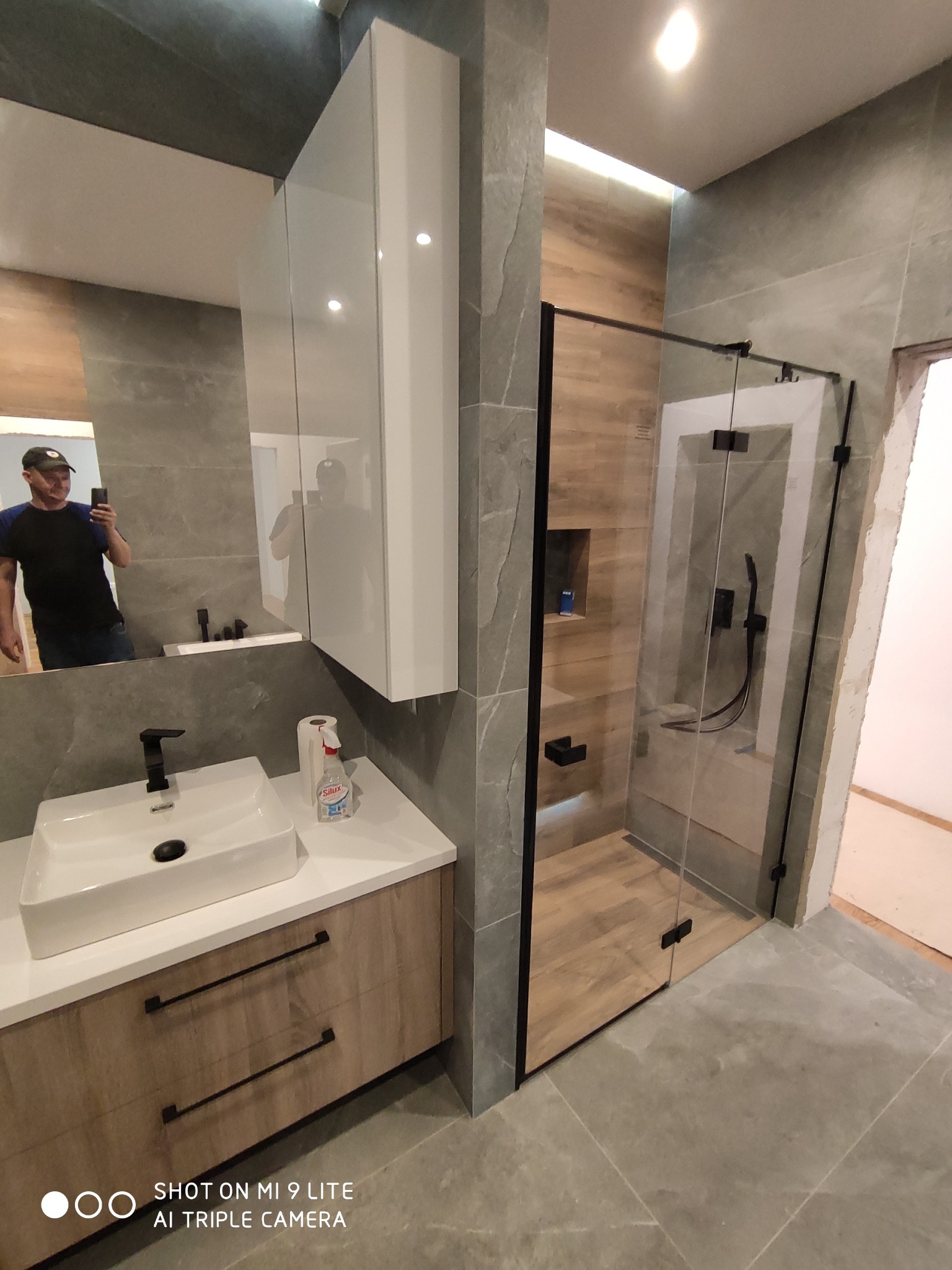 Nowoczesna łazienka po remoncie: kabina prysznicowa ze szklanymi drzwiami w czarnej ramie, umywalka nablatowa z czarną armaturą, biała szafka wisząca. W lustrze odbija się osoba robiąca zdjęcie.