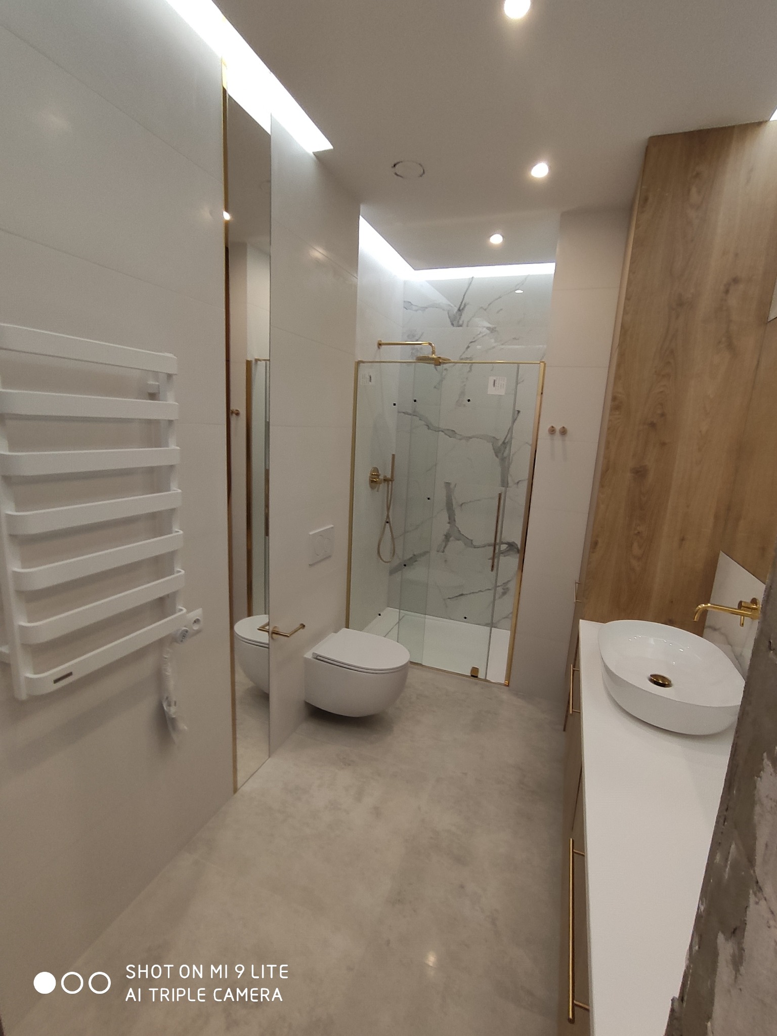 Nowoczesna łazienka z białymi płytkami, kabiną prysznicową ze złotymi ramami, podwieszaną toaletą i grzejnikiem drabinkowym, oświetlona ledami.