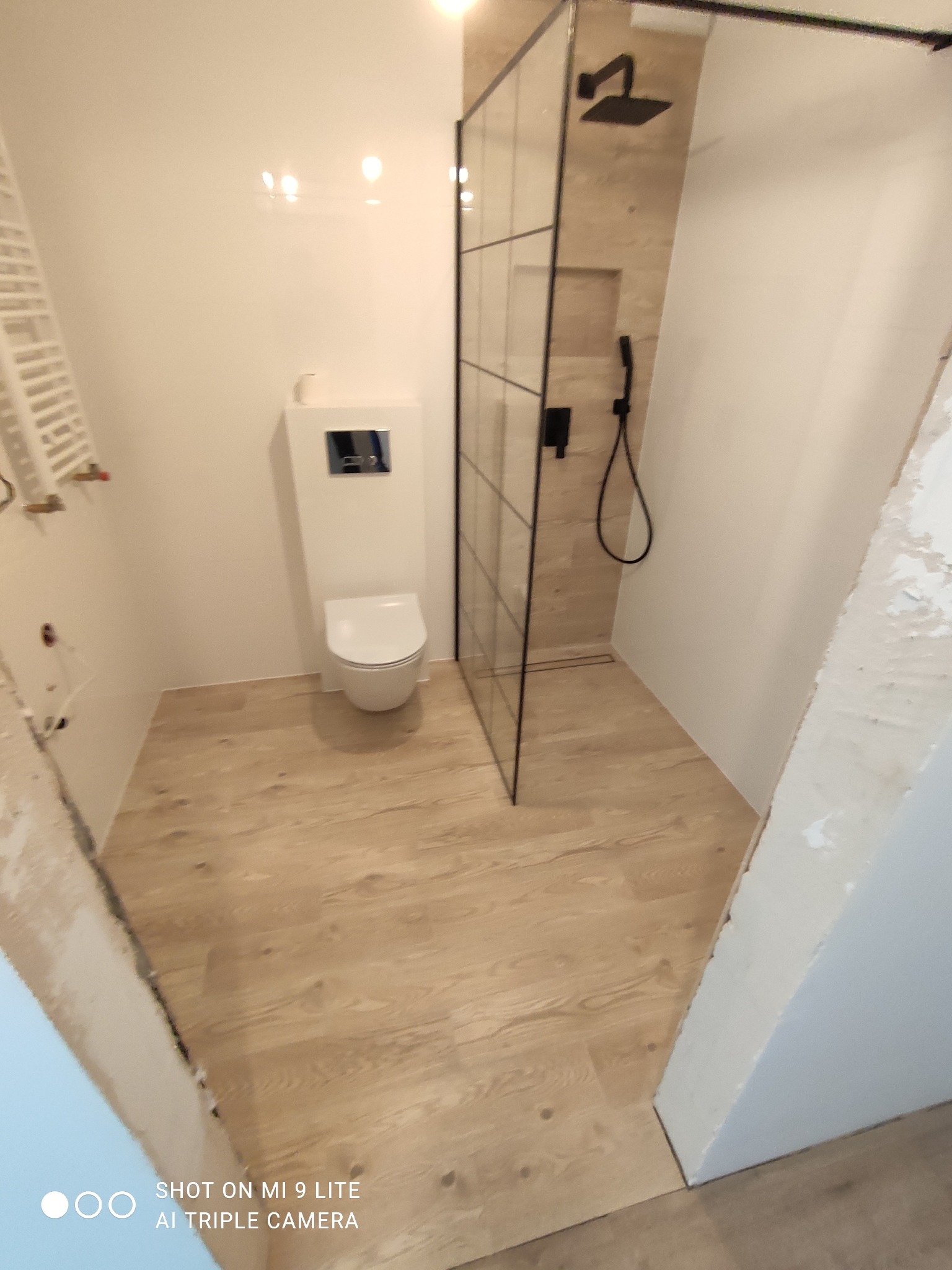 Nowoczesna łazienka w trakcie wykańczania: białe ściany, drewnopodobna podłoga, kabina prysznicowa z czarną armaturą i podtynkowy WC. Widoczne niedokończone obramowanie drzwi.