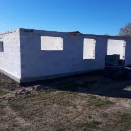 Surowy stan ścian zewnętrznych domu z betonu komórkowego, widoczne otwory okienne i drzwiowe, teren wokół w stanie naturalnym z piaskiem i trawą.