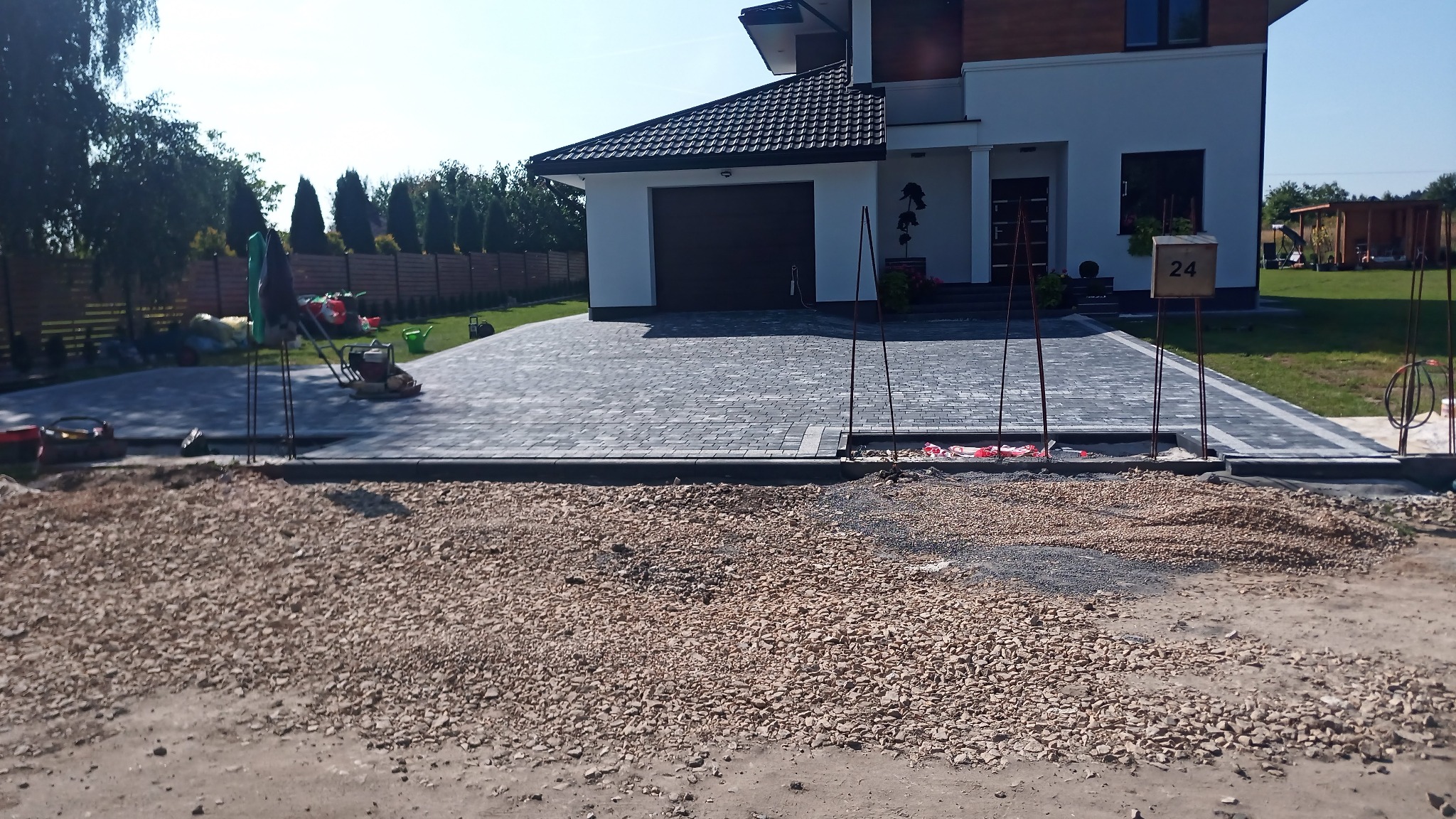 Nowa, szara kostka brukowa ułożona przed nowoczesnym domem z garażem, widoczne narzędzia i materiały budowlane, przygotowane podłoże z kamienia, numer domu 24.