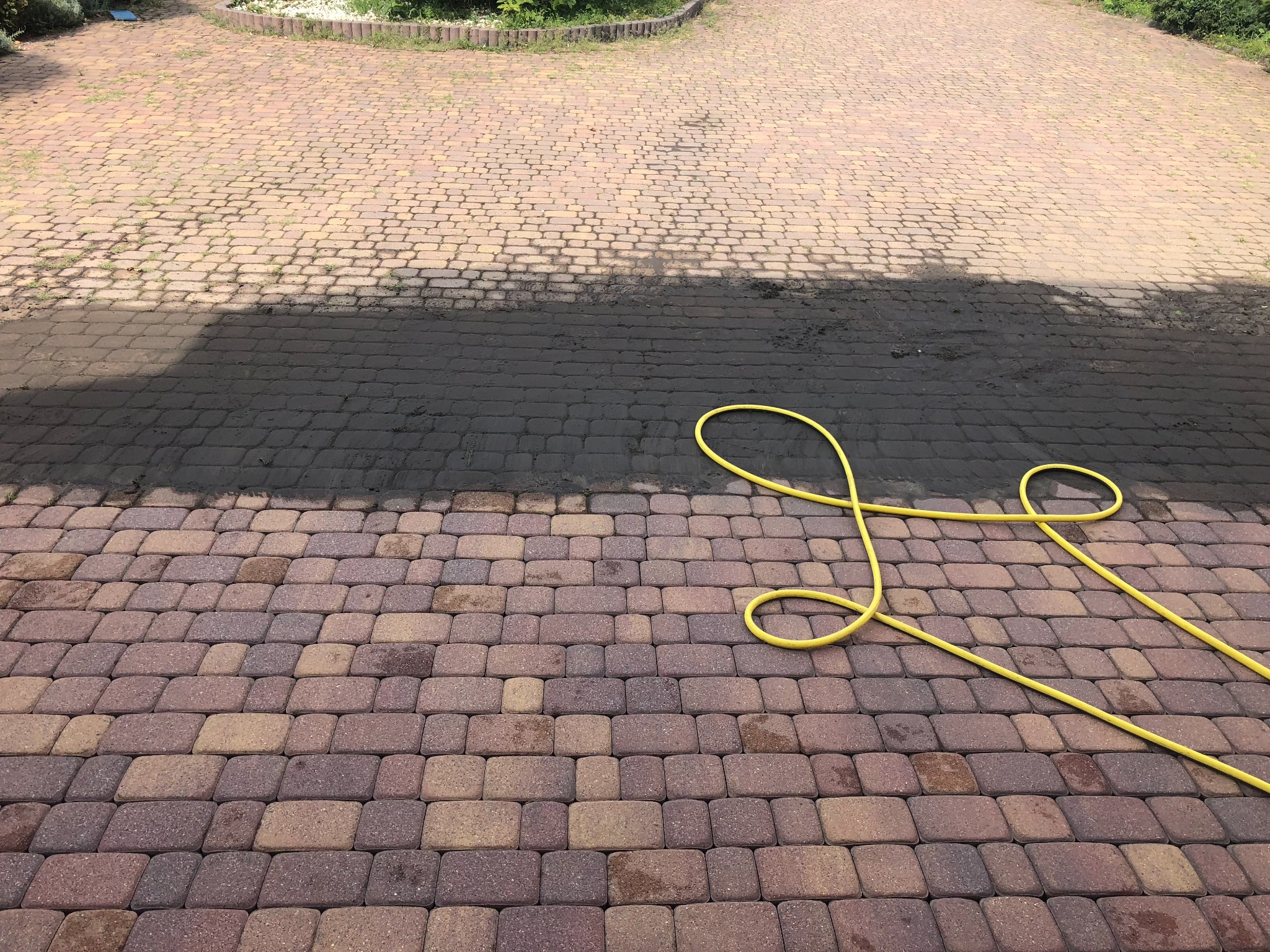 Częściowo oczyszczona, brudna kostka brukowa w odcieniach brązu i beżu, z żółtym wężem ogrodowym na wierzchu, z widocznym fragmentem trawnika w tle.