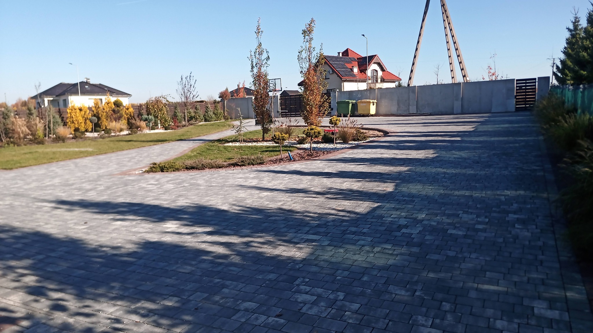 Szeroki podjazd z szarej kostki brukowej prowadzący do nowoczesnego domu z czerwonym dachem i panelami słonecznymi, widoczne elementy krajobrazu: trawnik, drzewa i krzewy ozdobne, a w tle betonowe...