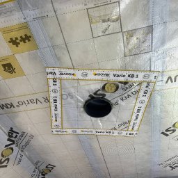 RN-BUD SERHII ZOSYK - Membrana paroizolacyjna Isover Vario KB 1 z wyciętym otworem na rurę, zabezpieczona taśmą mierniczą. Widoczne oznaczenia i siatka na membranie.