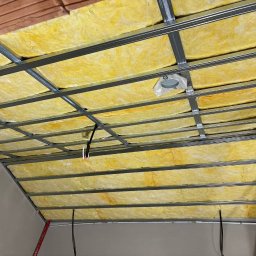 RN-BUD SERHII ZOSYK - Żółta wełna mineralna w metalowym stelażu sufitu podwieszanego, widok od dołu. Fragment ściany i instalacji elektrycznej.