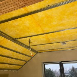 RN-BUD SERHII ZOSYK - Żółta wełna mineralna w metalowej konstrukcji sufitu poddasza, widoczny fragment okna dachowego i ściana. Ocieplenie izolacyjne na poddaszu.