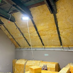 RN-BUD SERHII ZOSYK - Ocieplenie poddasza wełną mineralną w Bochni. Widoczne drewniane belki i halogen. Częściowo wykonana izolacja, materiały przygotowane do dalszych prac.