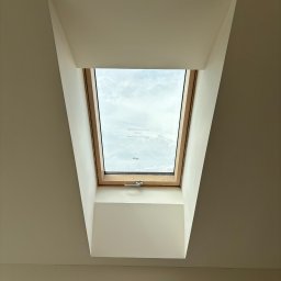 RN-BUD SERHII ZOSYK - Wykończone obramowanie okna dachowego na poddaszu z gładkimi ścianami i sufitem. Widok na pochmurne niebo przez okno.