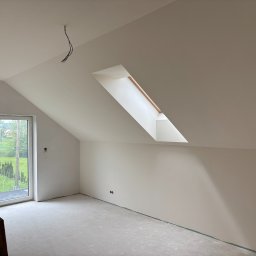 RN-BUD SERHII ZOSYK - Poddasze po gładziowaniu: białe ściany, okno dachowe, surowe podłoże. Widoczne kable oświetleniowe i otwór okienny z widokiem na zieleń. Minimalistyczne wykończenie wnętrza.