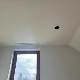 Poddasze po remoncie: świeżo pomalowane ściany w odcieniach szarości, okno dachowe z widokiem na zewnątrz, otwór na oświetlenie w suficie i kratka wentylacyjna.