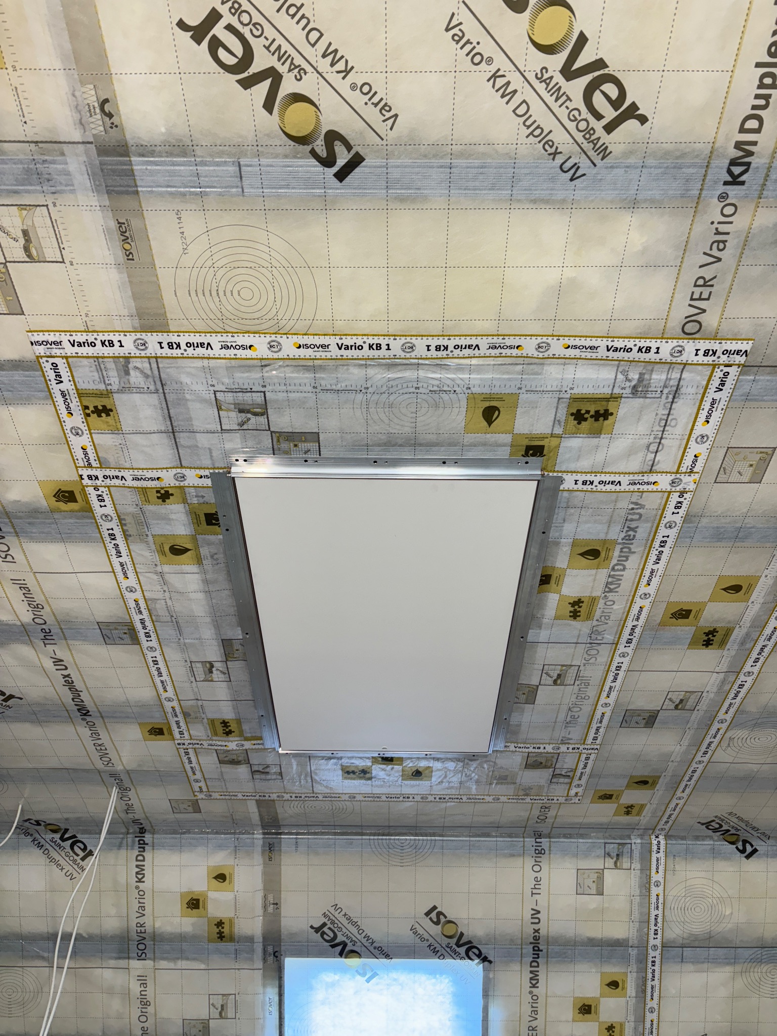 Montaż oświetlenia w suficie z izolacją Isover Vario KM Duplex UV, widoczne taśmy i materiały izolacyjne z nadrukowanymi oznaczeniami. Kąt ujęcia z dołu podkreśla trapezowy kształt otworu na lampę.