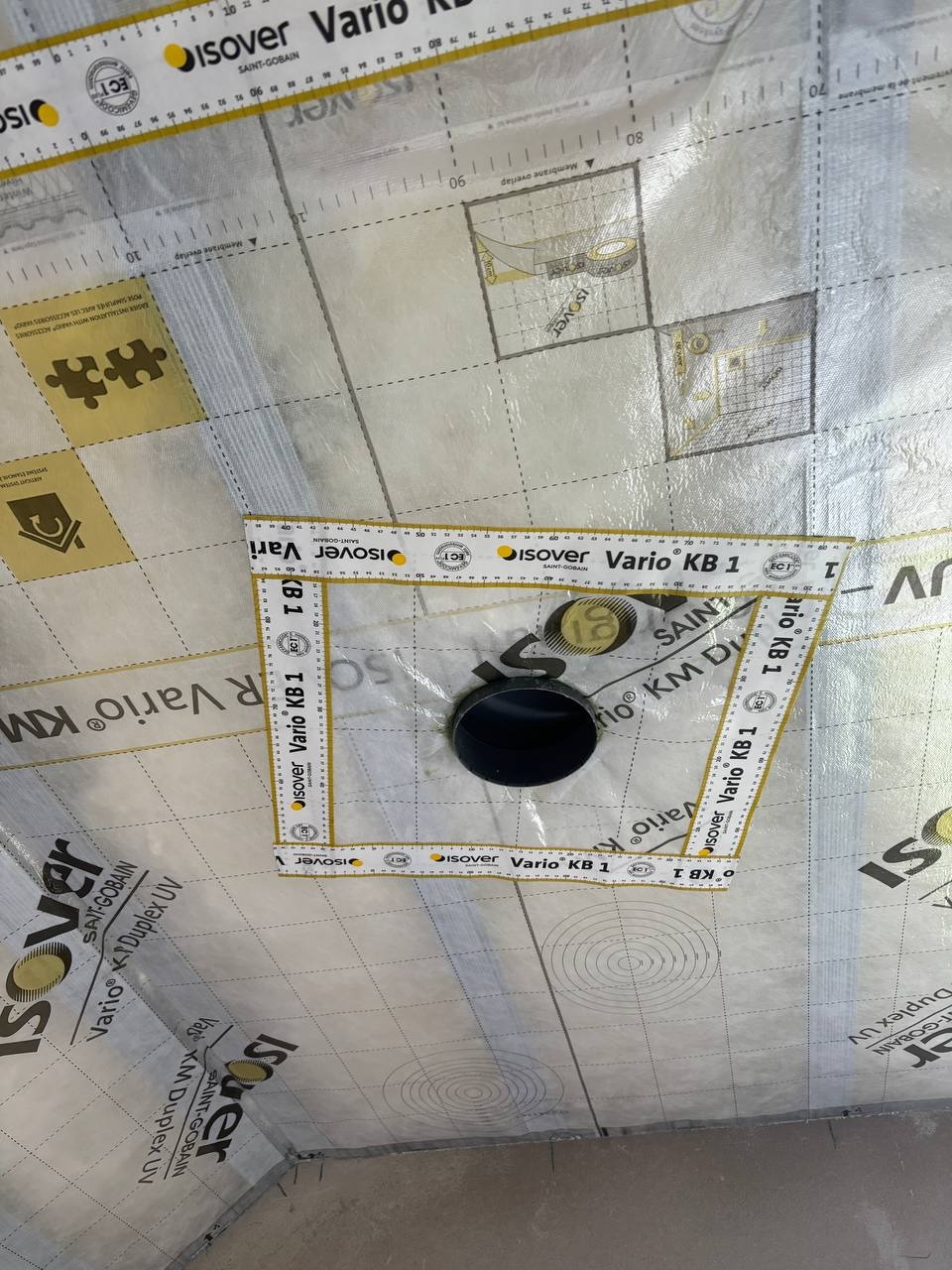 Membrana paroizolacyjna Isover Vario KB 1 z wyciętym otworem na rurę, zabezpieczona taśmą mierniczą. Widoczne oznaczenia i siatka na membranie.