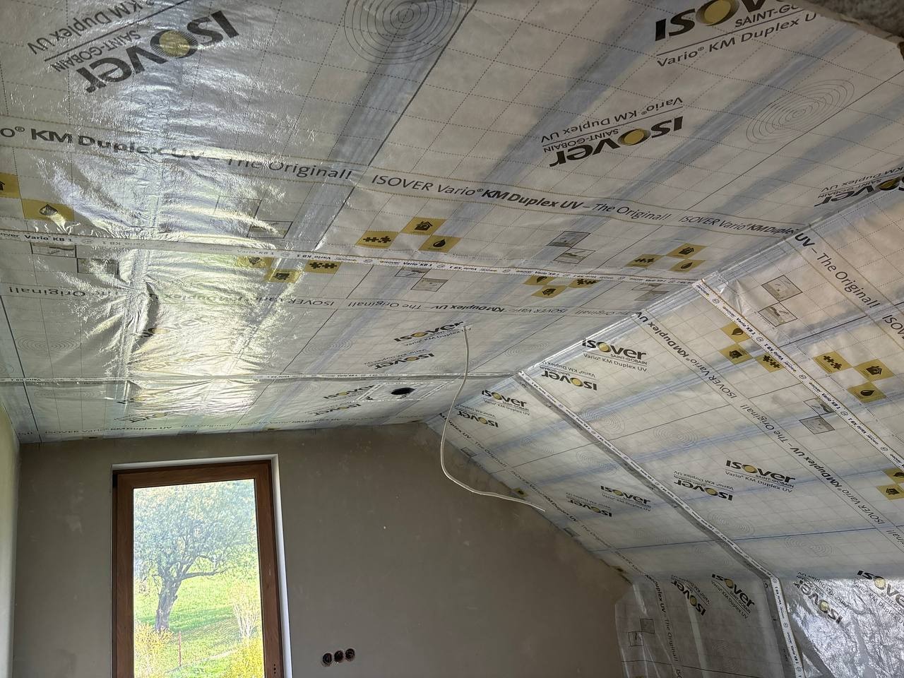 Ocieplenie poddasza folią paroizolacyjną Isover Vario KM Duplex UV, widok na okno i fragment ściany. Etap prac przed dalszym wykończeniem.