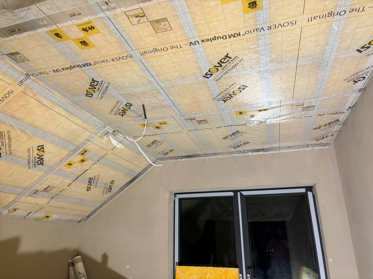 Izolacja poddasza folią paroizolacyjną Isover Vario KM Duplex UV. Widoczne elementy instalacji i okno. Prace wykończeniowe w jasnym pomieszczeniu.