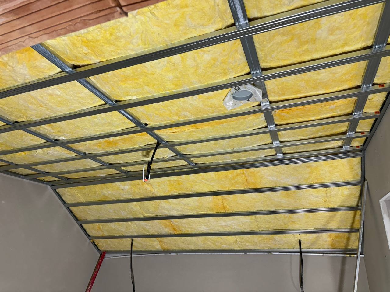 Żółta wełna mineralna w metalowym stelażu sufitu podwieszanego, widok od dołu. Fragment ściany i instalacji elektrycznej.