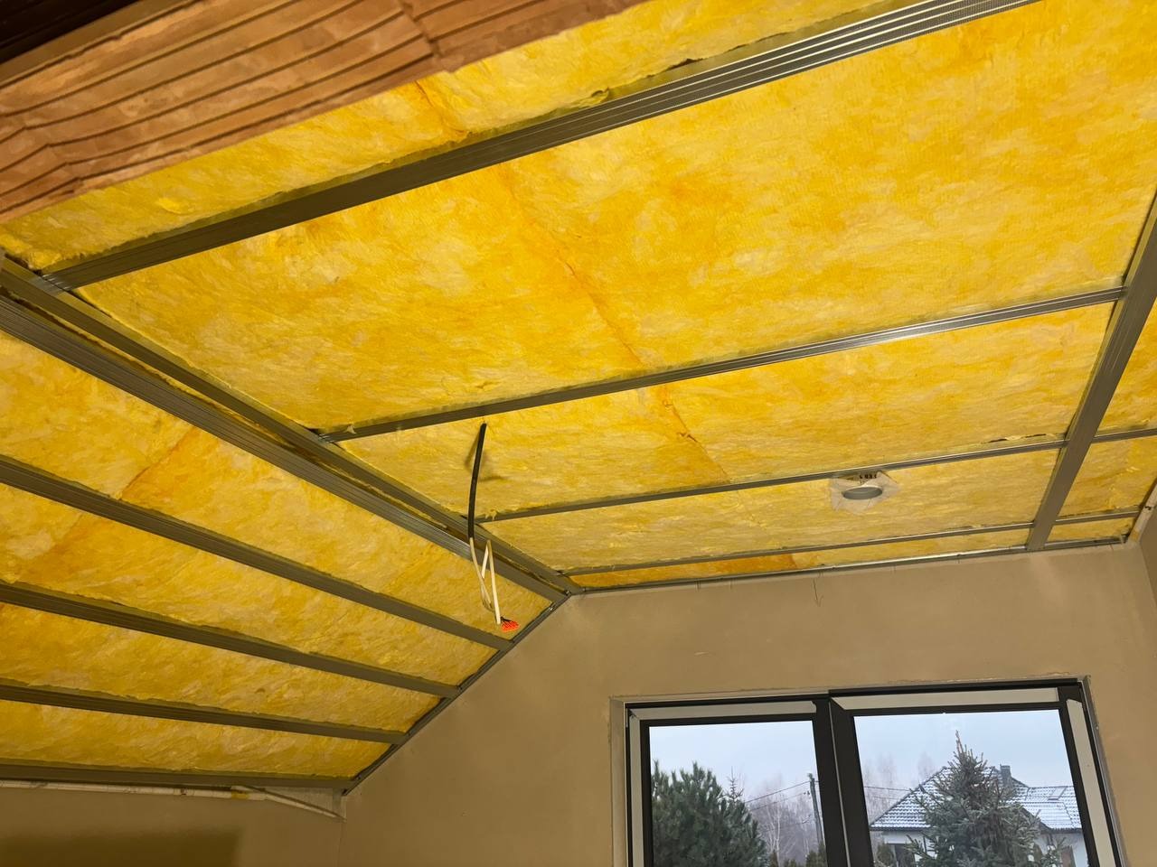 Żółta wełna mineralna w metalowej konstrukcji sufitu poddasza, widoczny fragment okna dachowego i ściana. Ocieplenie izolacyjne na poddaszu.