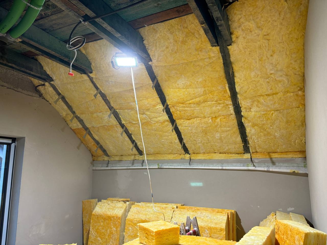 Ocieplenie poddasza wełną mineralną w Bochni. Widoczne drewniane belki i halogen. Częściowo wykonana izolacja, materiały przygotowane do dalszych prac.