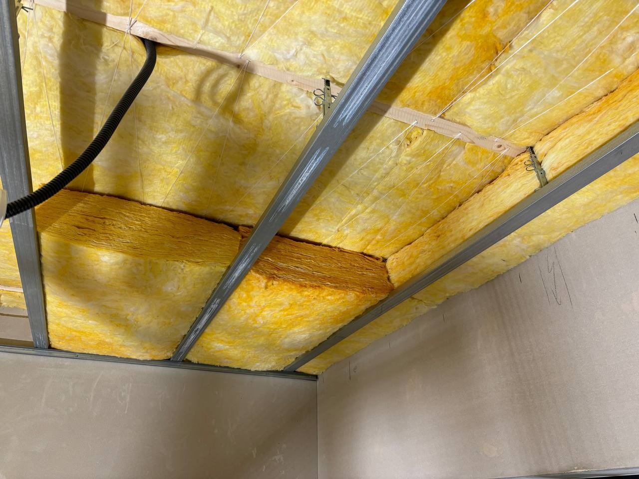 Poddasze ocieplone wełną mineralną, widoczne metalowe profile konstrukcyjne. Kabel elektryczny w peszlu w rogu. Ocieplenie w kolorze żółtym, świeżo położone.