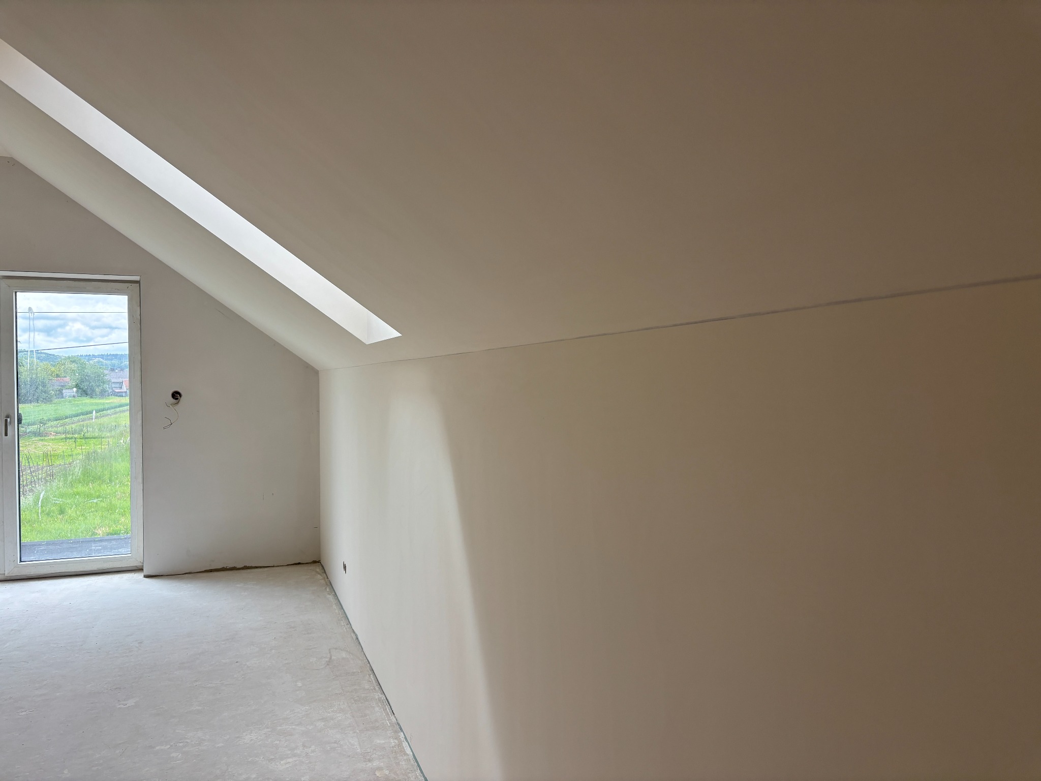 Poddasze po remoncie: gładkie, jasne ściany w odcieniach beżu, okno dachowe i widok na zieleń za oknem. Minimalistyczne wykończenie wnętrza.