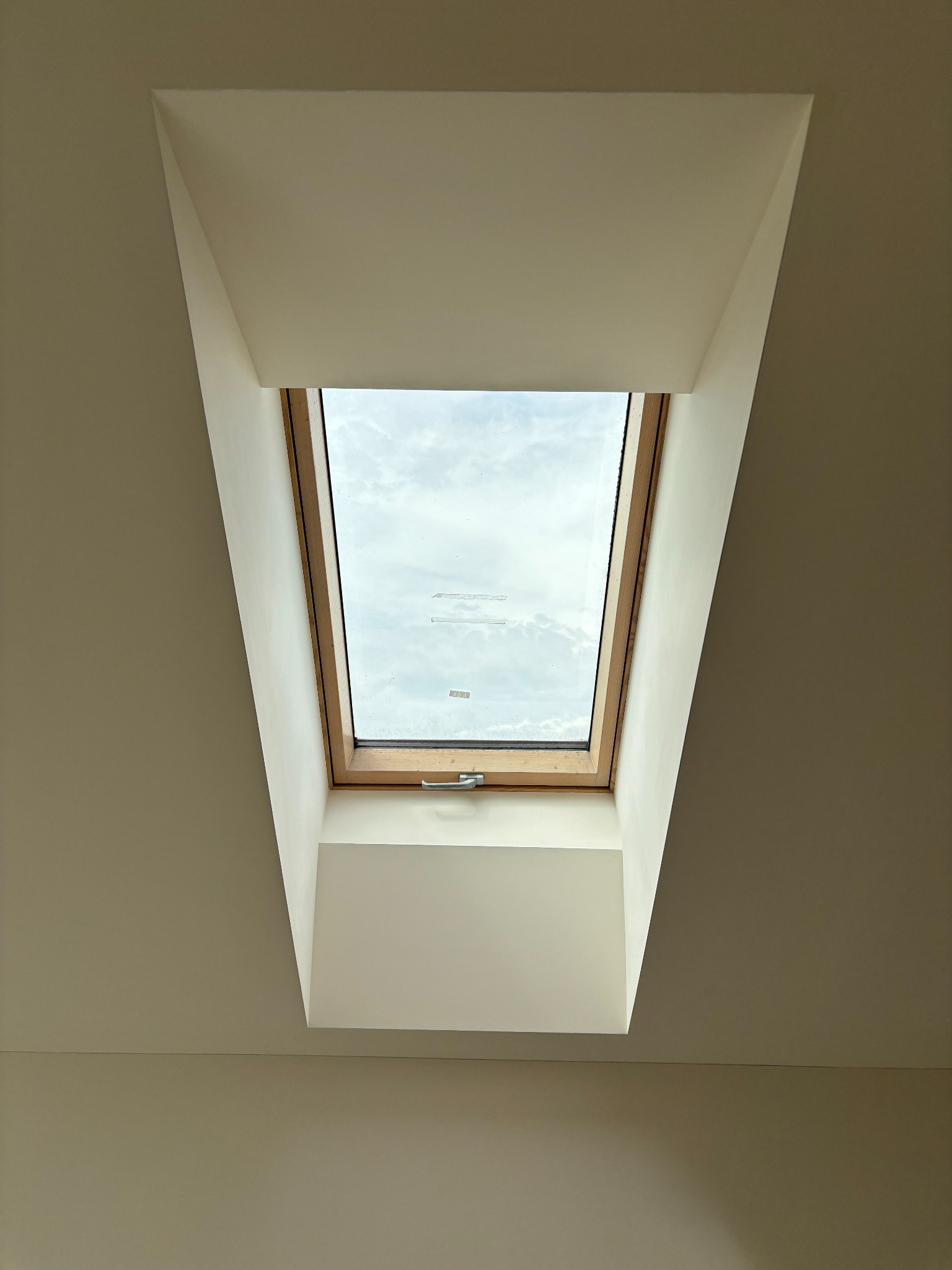 Wykończone obramowanie okna dachowego na poddaszu z gładkimi ścianami i sufitem. Widok na pochmurne niebo przez okno.