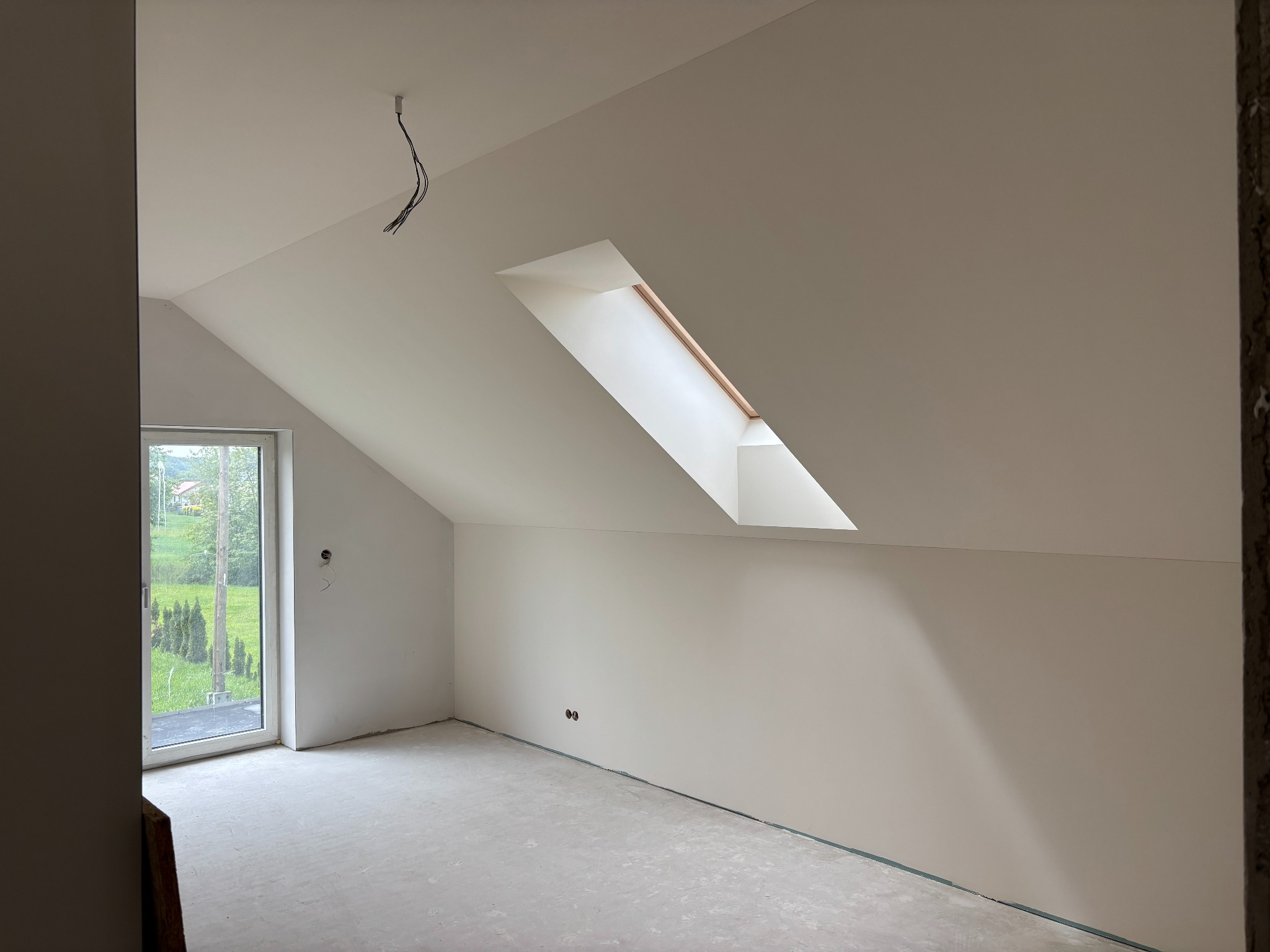 Poddasze po gładziowaniu: białe ściany, okno dachowe, surowe podłoże. Widoczne kable oświetleniowe i otwór okienny z widokiem na zieleń. Minimalistyczne wykończenie wnętrza.