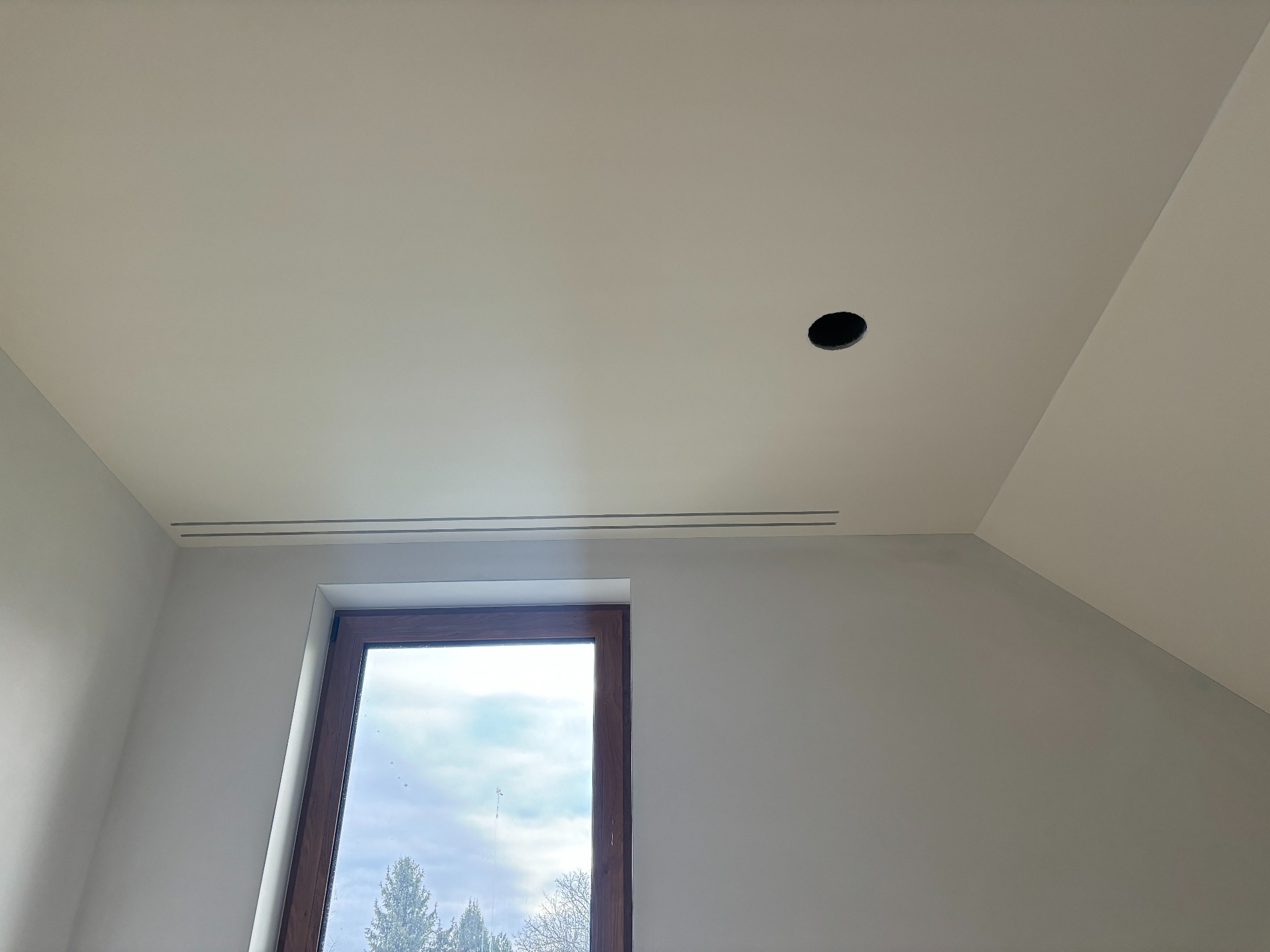 Poddasze po remoncie: świeżo pomalowane ściany w odcieniach szarości, okno dachowe z widokiem na zewnątrz, otwór na oświetlenie w suficie i kratka wentylacyjna.