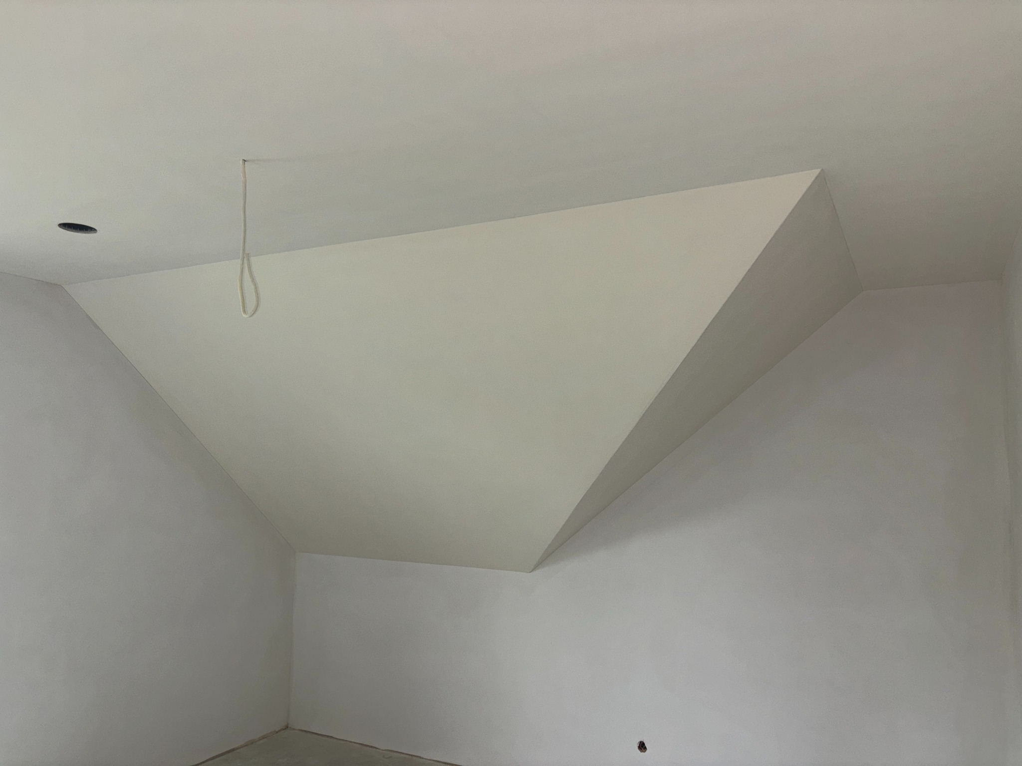 Poddasze po szpachlowaniu i malowaniu na biało. Widoczne skośne ściany i sufit, z otworem na oświetlenie i wystającym przewodem. Ściany gładkie, jasne, gotowe do dalszych prac.