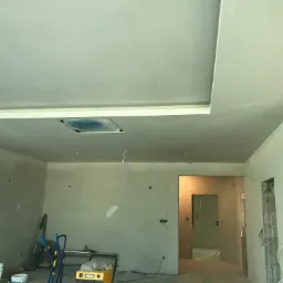 Wnętrze w trakcie prac wykończeniowych: sufit z gładzią, ściany w trakcie szpachlowania, widoczne narzędzia i materiały budowlane, wystające kable elektryczne.