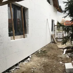 Biała elewacja budynku w trakcie ocieplania, widoczne płyty styropianowe i drewniane podpory okna, teren wokół budynku z resztkami materiałów izolacyjnych i ziemią.