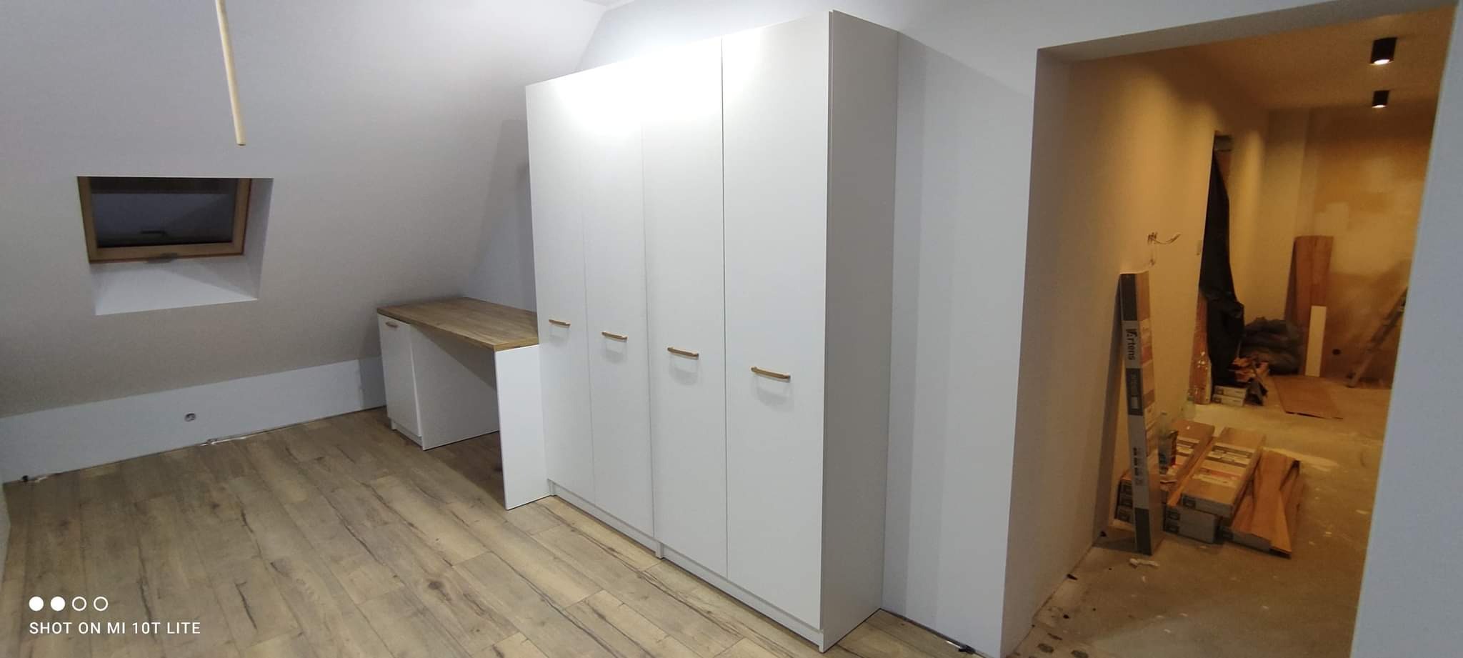 Biała szafa z czterema drzwiami i drewnianymi uchwytami, zintegrowana z biurkiem o drewnianym blacie, w pokoju na poddaszu z oknem dachowym; w tle widoczne pomieszczenie w trakcie remontu.