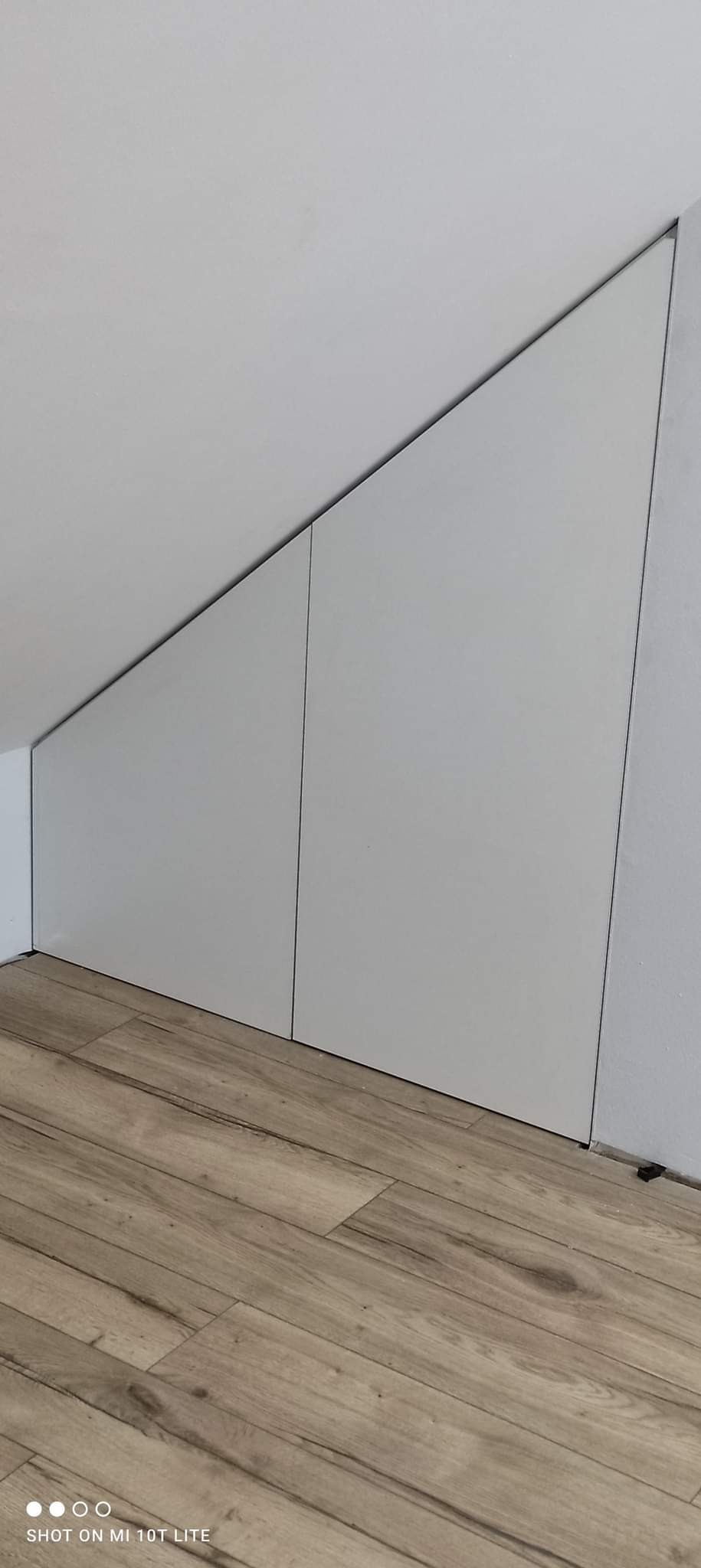 Biała szafa wnękowa z dwiema pionowymi sekcjami, dopasowana do skosu sufitu, na podłodze z paneli drewnopodobnych.