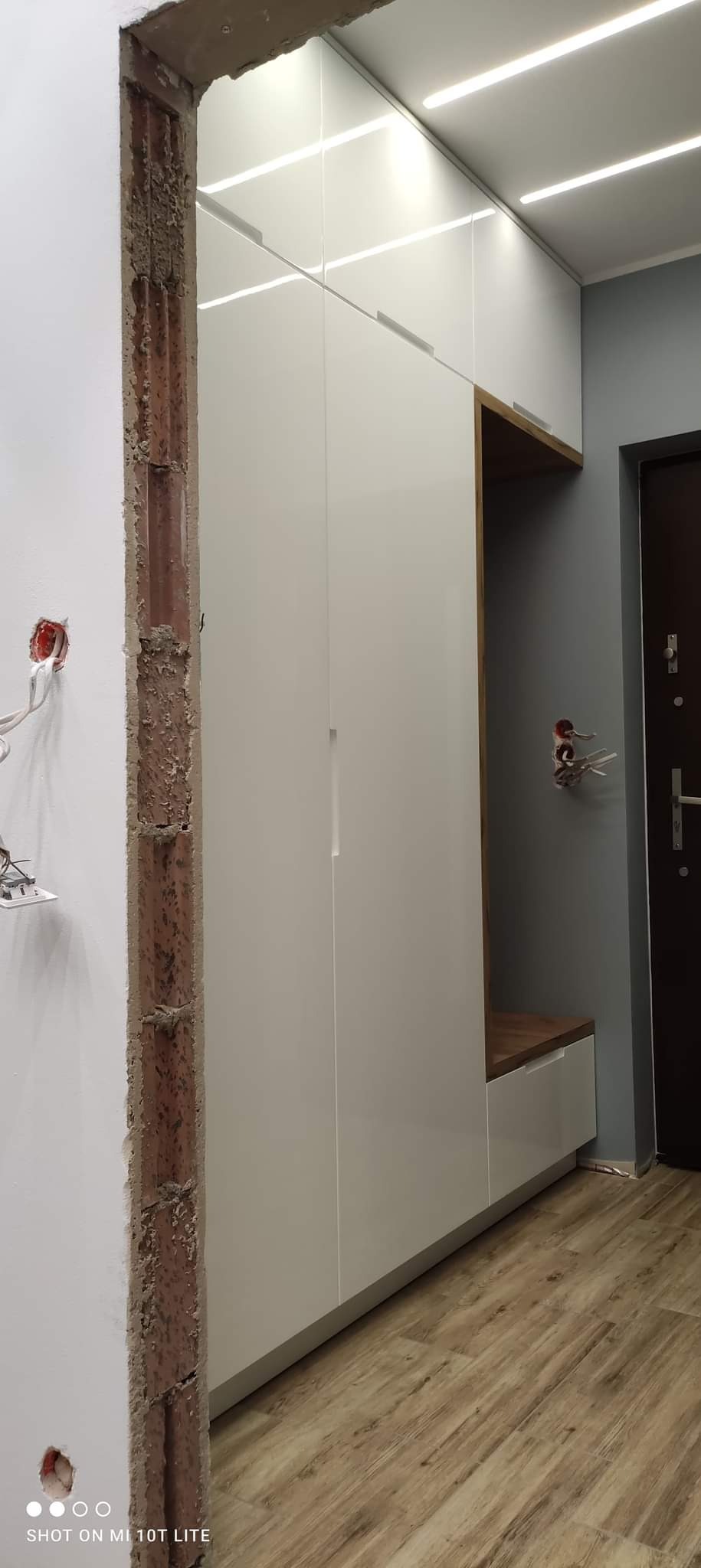 Biała szafa wnękowa z drewnianymi akcentami i siedziskiem w Ziębicach, widoczna podczas montażu z odsłoniętą cegłą w ścianie.