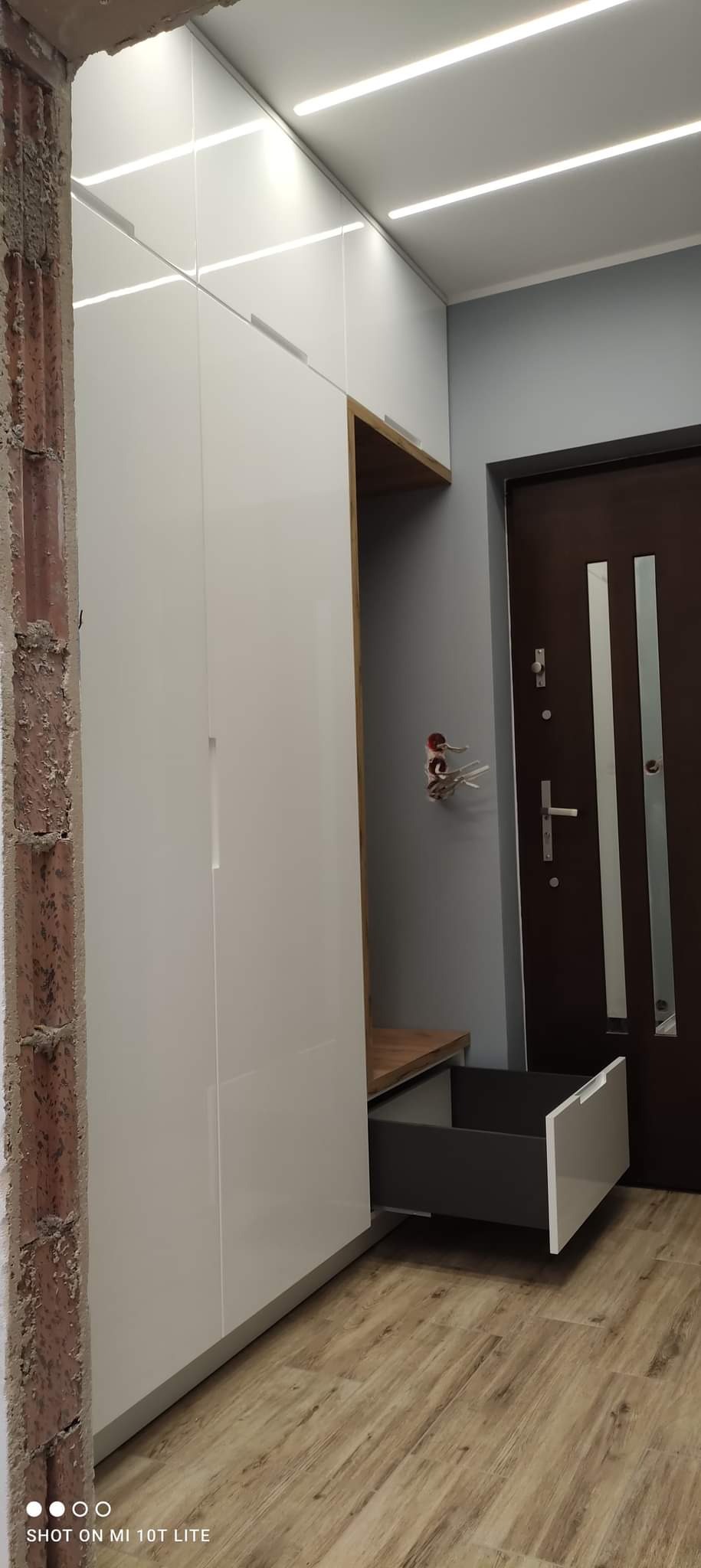 Biała szafa wnękowa z wysuwaną szufladą, widoczny fragment ściany w stanie surowym i drewnopodobna podłoga.