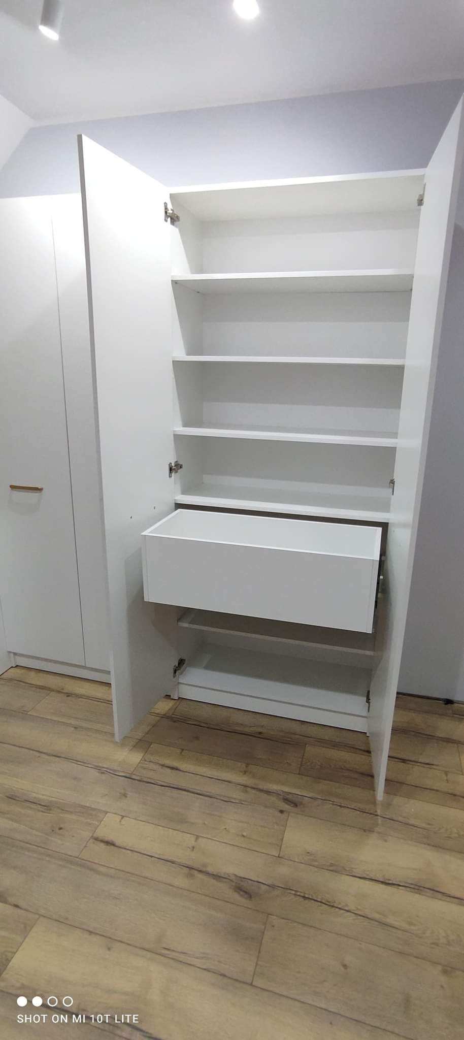 Biała szafa wnękowa z otwartymi drzwiami, widoczne półki i szuflada, drewniana podłoga, minimalistyczny design.