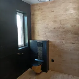 Nowoczesna łazienka z kontrastującymi czarnymi i drewnopodobnymi płytkami, wisząca toaleta z drewnianą obudową, minimalistyczne oświetlenie sufitowe.