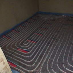 Daw-Instal - Rozłożone rury ogrzewania podłogowego na izolacji z folią aluminiową, zabezpieczone taśmą brzegową przy ścianie, widoczne czarne klipsy mocujące rury do podłoża.