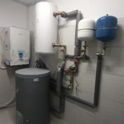 Kotłownia w trakcie instalacji: biały zbiornik ciepłej wody owinięty folią ochronną, szary zasobnik, niebieski zbiornik przeponowy oraz biały piec gazowy Panasonic, widoczne rury instalacyjne...