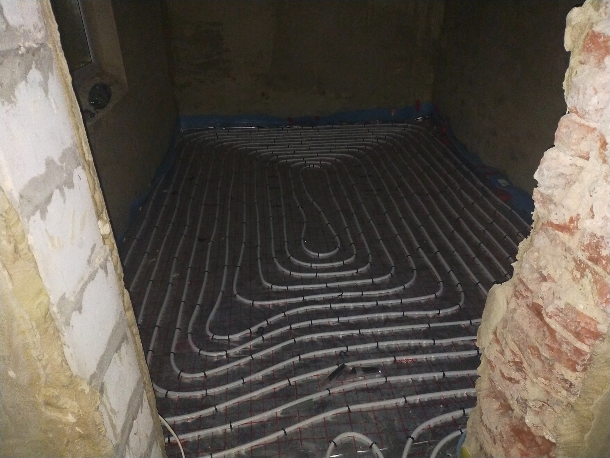 Rozłożone rury ogrzewania podłogowego na siatce w surowym pomieszczeniu z fragmentami muru i tynku.