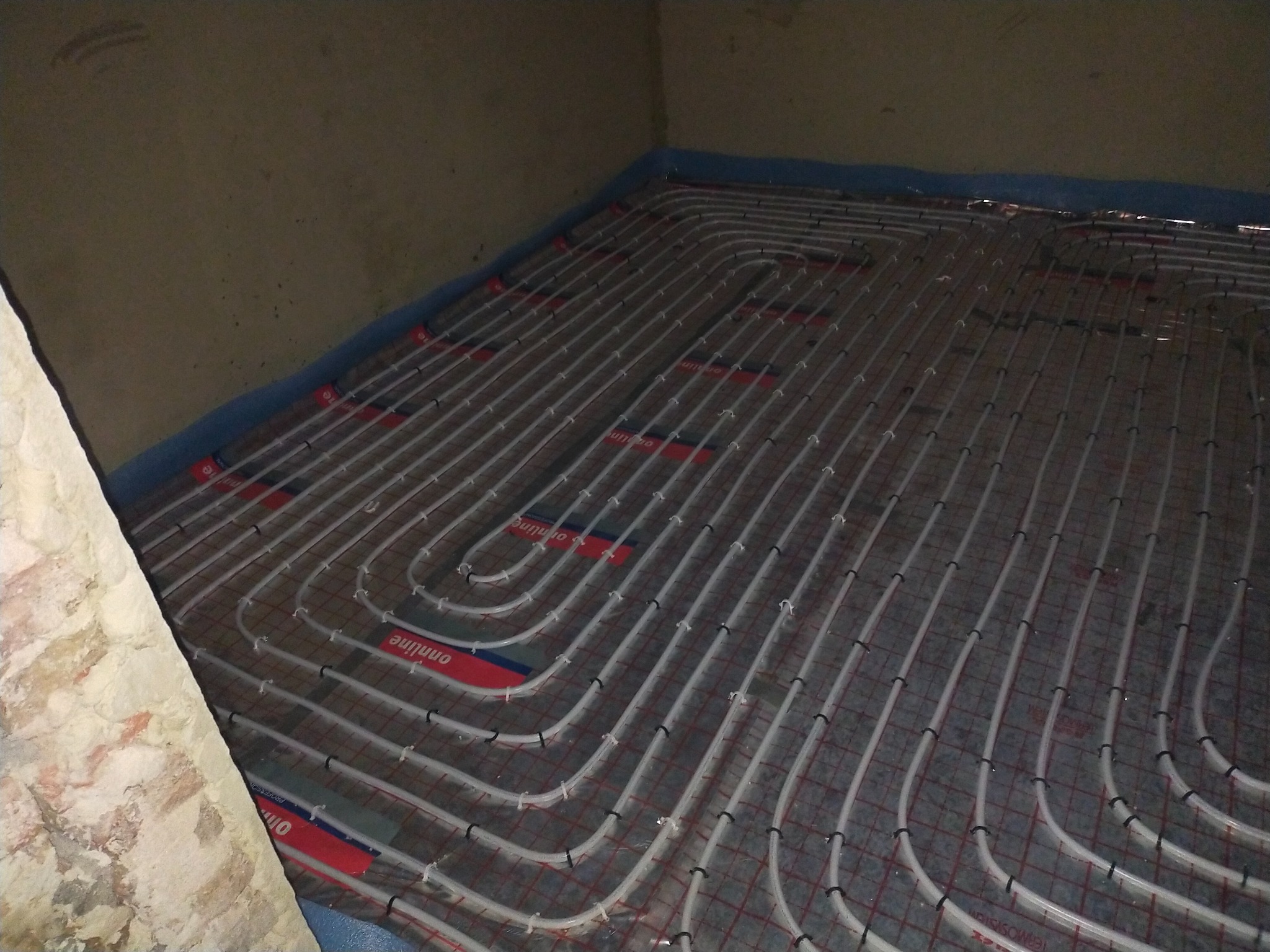 Rozłożone rury ogrzewania podłogowego na izolacji z folią aluminiową, zabezpieczone taśmą brzegową przy ścianie, widoczne czarne klipsy mocujące rury do podłoża.