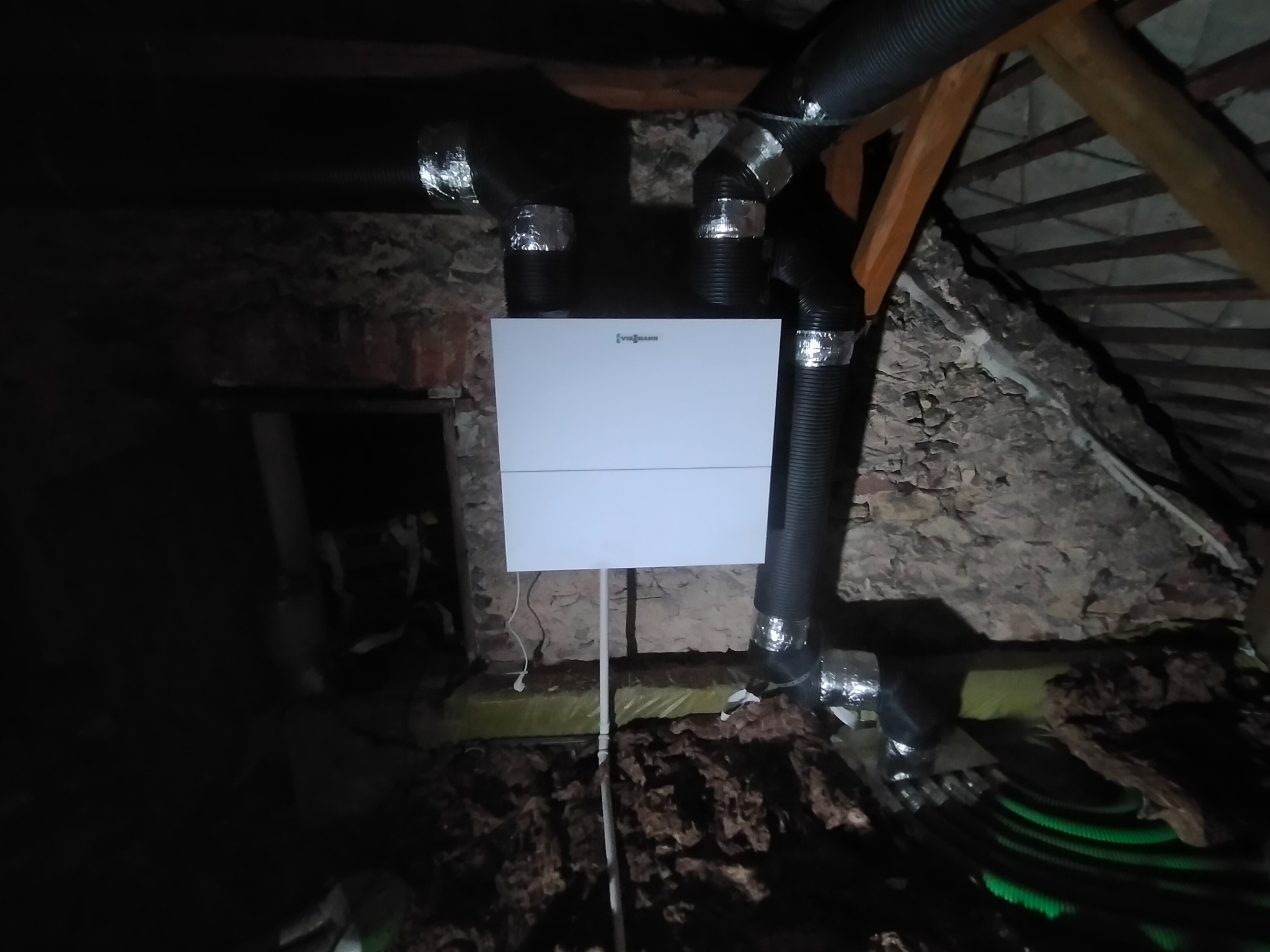 Biały piec Viessmann z czarnymi przewodami wentylacyjnymi w folii aluminiowej, zamontowany na poddaszu o kamiennych ścianach i drewnianej konstrukcji dachu.