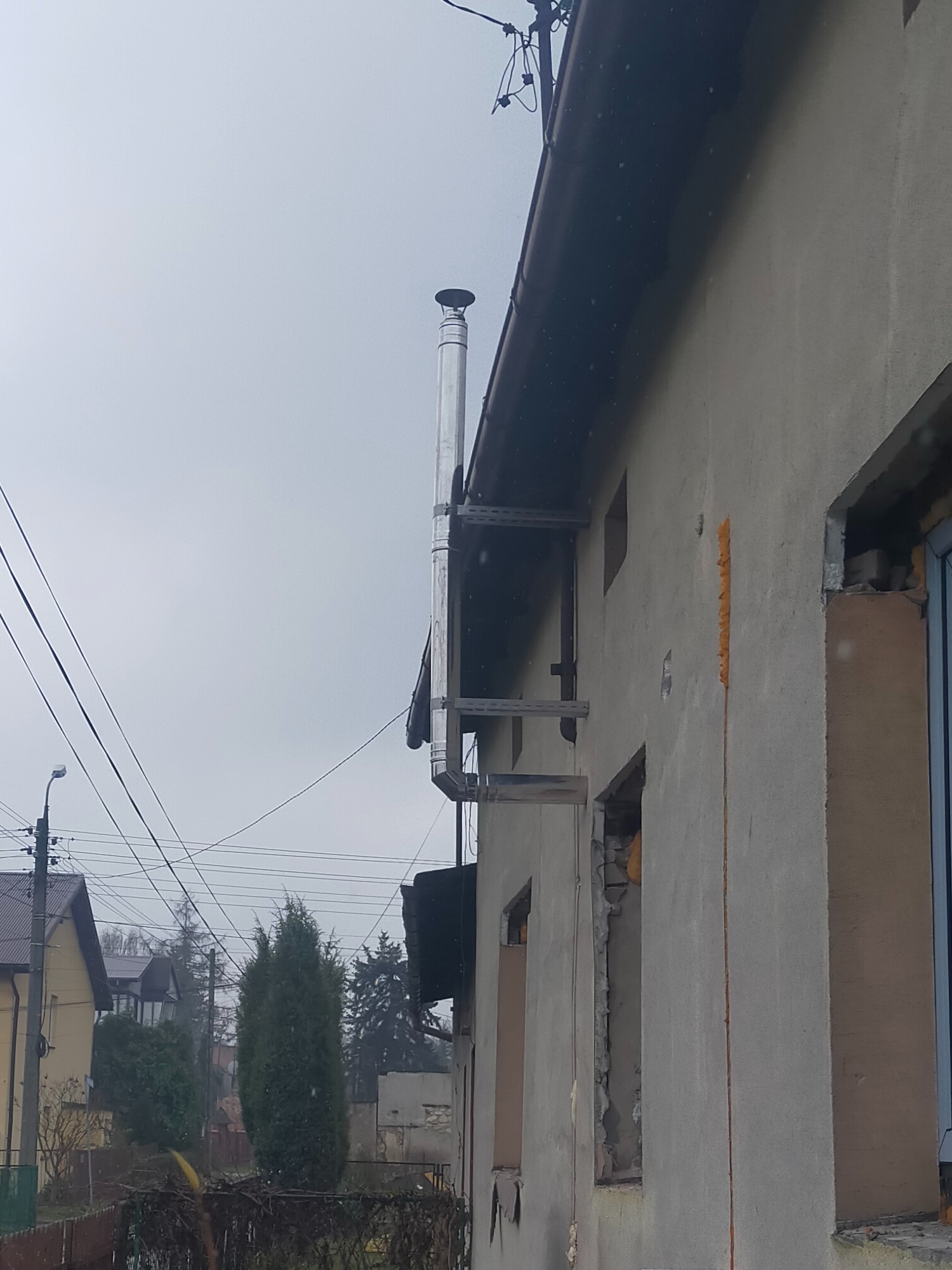 Zewnętrzny montaż stalowego komina spalinowego przy ścianie budynku z widocznymi elementami konstrukcyjnymi wsparcia, w tle zabudowa jednorodzinna i pochmurne niebo.