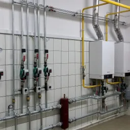 Dwa białe kotły grzewcze Viessmann zamontowane na ścianie z białych płytek, z widocznymi rurami gazowymi (żółte) i wodnymi (srebrne), zaworami, manometrami oraz czerwonymi regulatorami na ścianie.