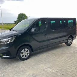 Czarny, długi bus marki Renault Trafic na brukowanym placu, z widocznym fragmentem zieleni w tle.