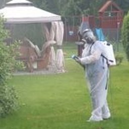 Ozonlux - Osoba w białym kombinezonie ochronnym, z plecakiem opryskiwaczem, rozpyla substancję na trawniku przed domem w słoneczny dzień. Widoczna mgiełka oprysku.