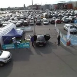 Stoisko promocyjne na parkingu przed centrum handlowym, z rozstawionym namiotem i samochodem z otwartymi drzwiami, w tle zaparkowane liczne pojazdy i budynek sklepu.