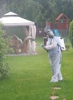 Osoba w białym kombinezonie ochronnym, z plecakiem opryskiwaczem, rozpyla substancję na trawniku przed domem w słoneczny dzień. Widoczna mgiełka oprysku.