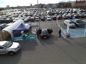 Stoisko promocyjne na parkingu przed centrum handlowym, z rozstawionym namiotem i samochodem z otwartymi drzwiami, w tle zaparkowane liczne pojazdy i budynek sklepu.