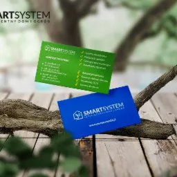 Dwie wizytówki firmy SmartSystem leżące na kawałku drewna na tle drewnianego stołu i zieleni. Jedna wizytówka jest zielona, druga niebieska. Na obu widoczne logo firmy i dane kontaktowe.