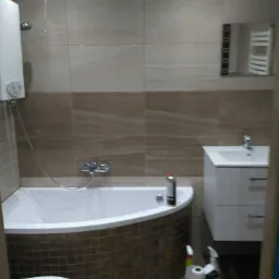 Wykończona łazienka z wanną obudowaną brązową mozaiką, białą umywalką z szafką i grzejnikiem drabinkowym w tle. Na wannie stoi puszka z preparatem, a na podłodze leżą środki czystości i paczka...